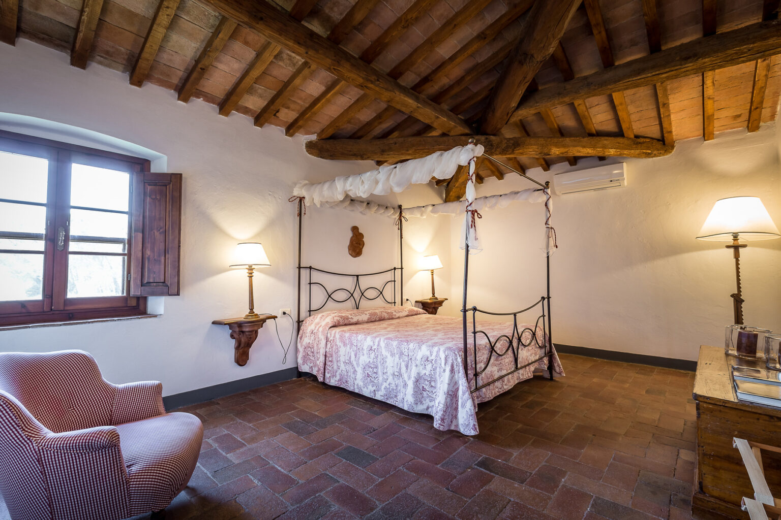 Cozy bedroom featuring a four-poster bed with a floral duvet, wooden beams on the ceiling, and a charming armchair. The room is illuminated by bedside lamps and has a window with wooden shutters, creating a warm and inviting atmosphere. | Only on SopranoVillas