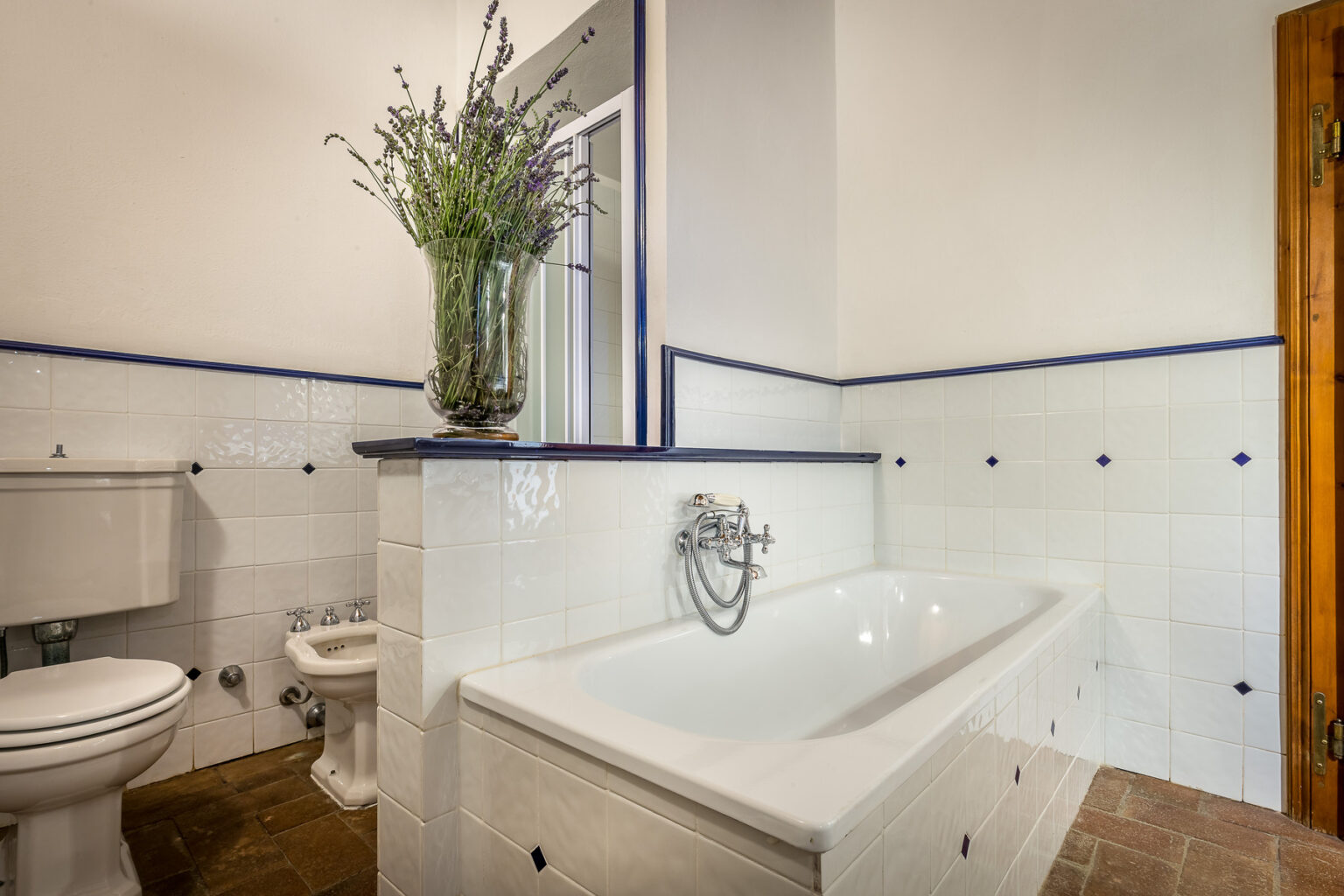 Spacious bathroom featuring a white bathtub, modern fixtures, and a decorative vase with lavender. The room is adorned with white tiles and blue accents, providing a clean and elegant atmosphere. | Only on SopranoVillas