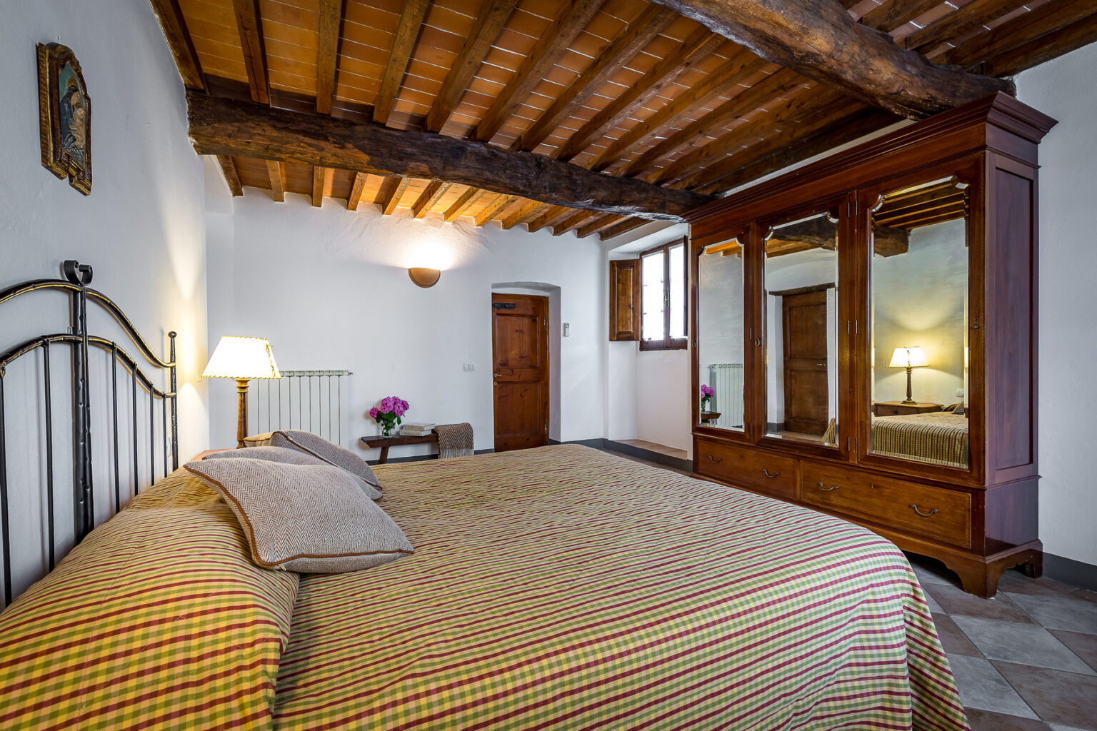 Cozy bedroom featuring a wrought iron bed with striped bedding, wooden ceiling beams, and a vintage wardrobe. The room includes a bedside lamp, a small table with flowers, and natural light from windows, creating a warm and inviting atmosphere. Ideal for a relaxing getaway. | Only on SopranoVillas