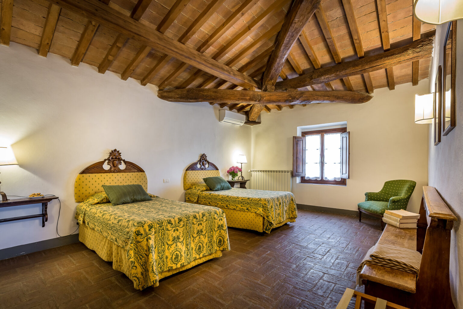 Cozy guest room featuring two twin beds with yellow floral bedspreads, wooden beamed ceiling, and a green armchair. The room includes a bedside lamp, a wooden bench, and a window with traditional shutters, creating a warm and inviting atmosphere. Ideal for travelers seeking comfort and style. | Only on SopranoVillas