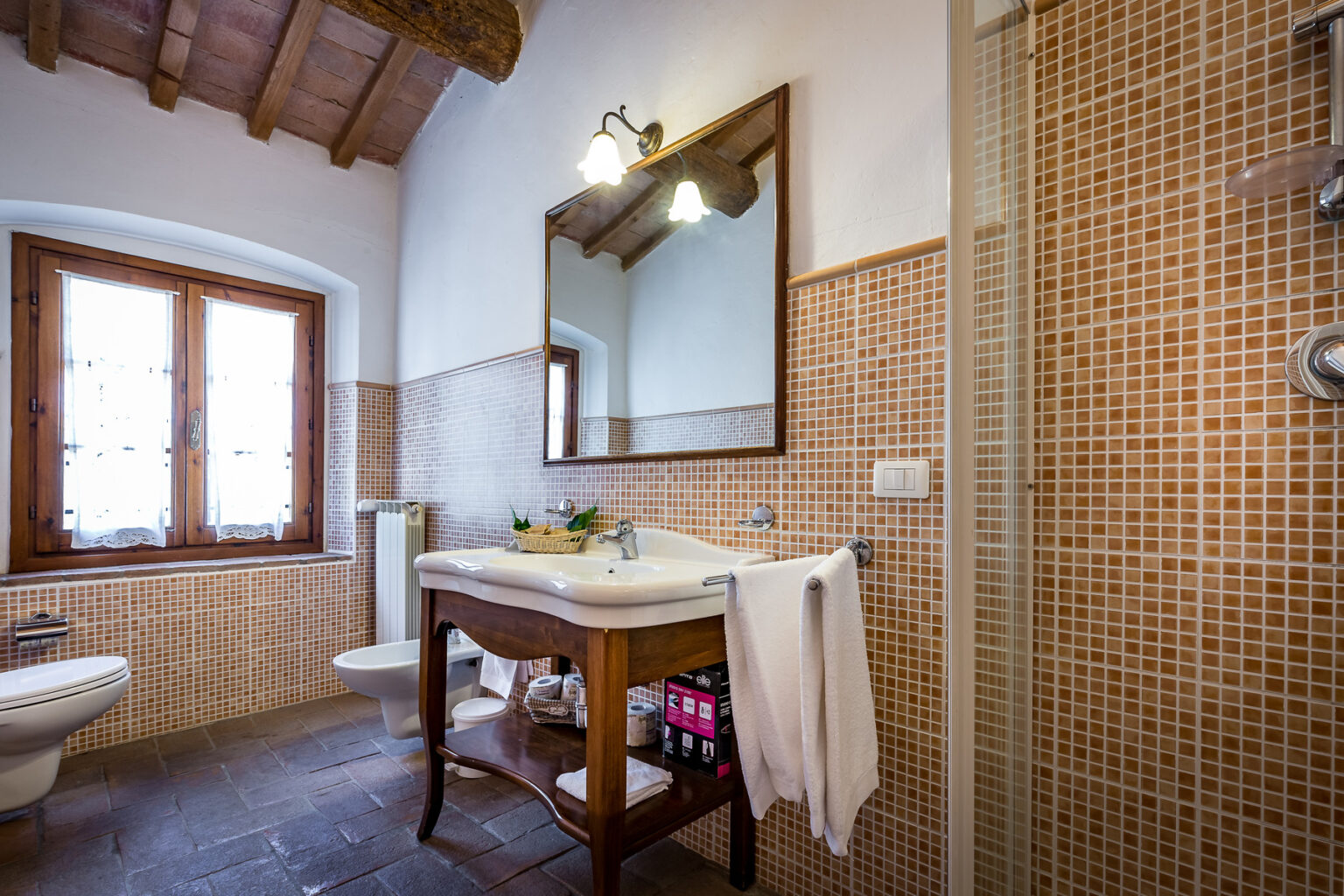 Cozy bathroom featuring a wooden ceiling, mosaic tile walls, a large mirror above a vintage sink, and natural light from two windows. The space includes a shower, toilet, and neatly arranged toiletries, creating a warm and inviting atmosphere. | Only on SopranoVillas