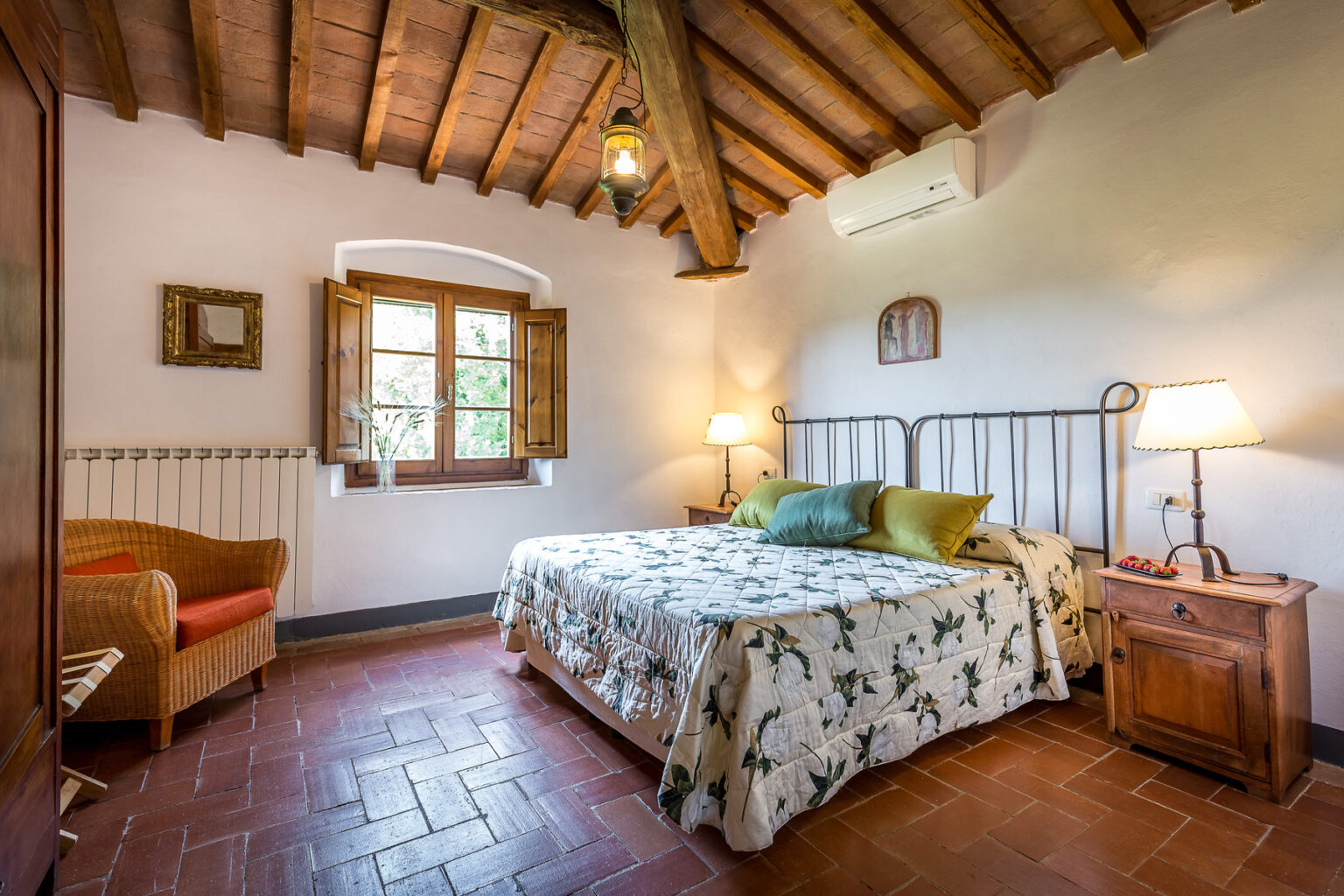 Cozy bedroom featuring a metal bed with floral bedding, wooden beams on the ceiling, and warm lighting. The room includes a comfortable armchair, bedside table, and a window with scenic views, creating a welcoming atmosphere. | Only on SopranoVillas
