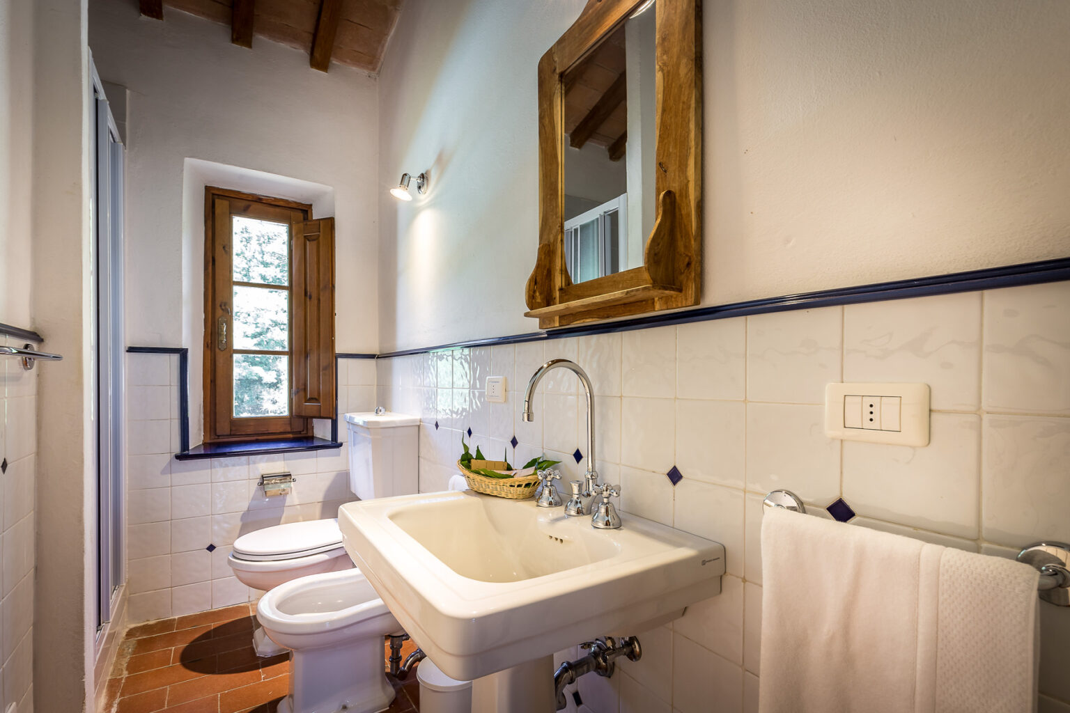 Bright and airy bathroom featuring a vintage wooden mirror, white sink, and modern toilet. Natural light streams in through a window, highlighting the tiled walls and wooden accents. Ideal for showcasing home decor and renovation ideas. | Only on SopranoVillas