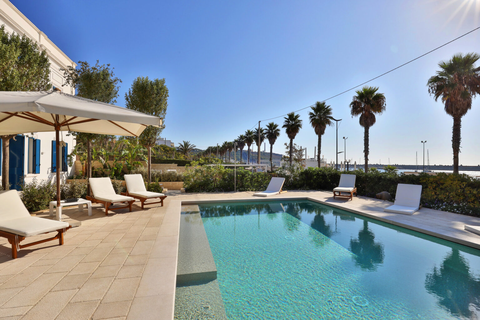 Luxurious outdoor pool area featuring sun loungers and an umbrella, surrounded by lush greenery and palm trees, with a clear blue sky and a glimpse of a waterfront in the background. Perfect for relaxation and leisure in a serene setting. | Only on SopranoVillas
