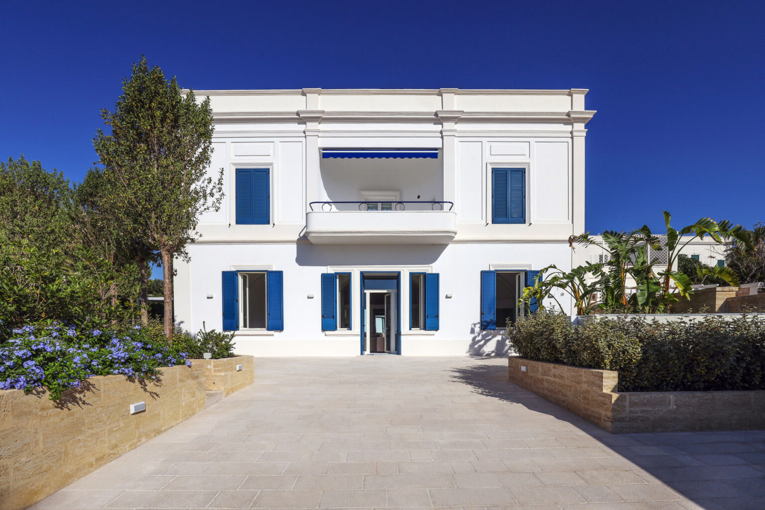 Modern white villa with blue shutters, surrounded by greenery and flowers, featuring a spacious stone pathway and clear blue sky. Ideal for luxury real estate listings or vacation rentals. | Only on SopranoVillas