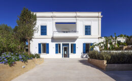 Modern white villa with blue shutters, surrounded by greenery and flowers, featuring a spacious stone pathway and clear blue sky. Ideal for luxury real estate listings or vacation rentals.