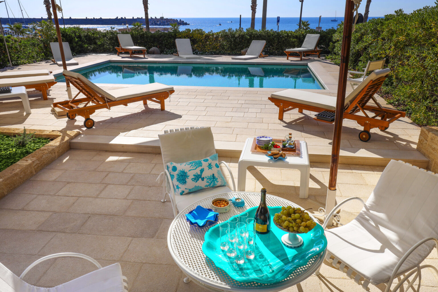 Luxurious outdoor pool area with comfortable lounge chairs, a table set with a bottle of champagne, glasses, and a bowl of grapes, overlooking a scenic ocean view. Perfect for relaxation and entertaining in a sunny, tropical setting. | Only on SopranoVillas