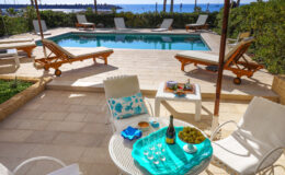 Luxurious outdoor pool area with comfortable lounge chairs, a table set with a bottle of champagne, glasses, and a bowl of grapes, overlooking a scenic ocean view. Perfect for relaxation and entertaining in a sunny, tropical setting.