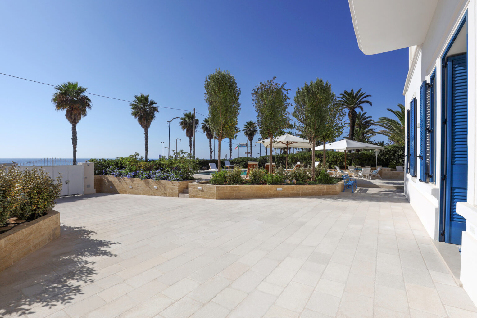 Coastal villa exterior featuring palm trees, landscaped gardens, and a spacious patio area with sun loungers and umbrellas, overlooking the sea under a clear blue sky. | Only on SopranoVillas
