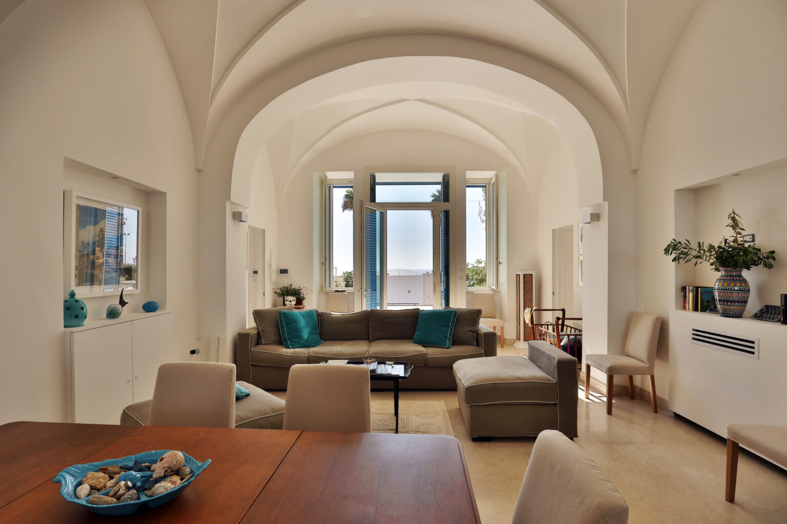 Spacious modern living room with high arched ceilings, featuring a comfortable beige sofa with turquoise cushions, a wooden dining table, and large windows allowing natural light. Decorative elements include a blue vase and a bowl of seashells on the table, creating a serene and inviting atmosphere. | Only on SopranoVillas