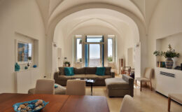 Spacious modern living room with high arched ceilings, featuring a comfortable beige sofa with turquoise cushions, a wooden dining table, and large windows allowing natural light. Decorative elements include a blue vase and a bowl of seashells on the table, creating a serene and inviting atmosphere.