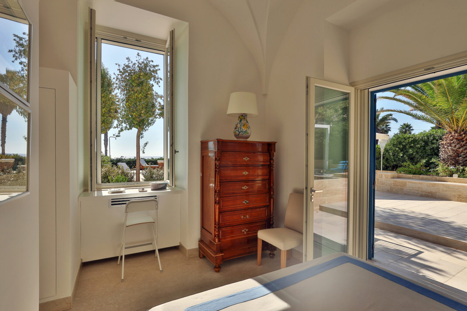 Bright and airy bedroom featuring a wooden dresser, a chair, and a small desk by the window. The window offers a view of palm trees and outdoor seating, enhancing the room's natural light and serene ambiance. | Only on SopranoVillas