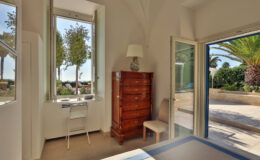 Bright and airy bedroom featuring a wooden dresser, a chair, and a small desk by the window. The window offers a view of palm trees and outdoor seating, enhancing the room's natural light and serene ambiance.