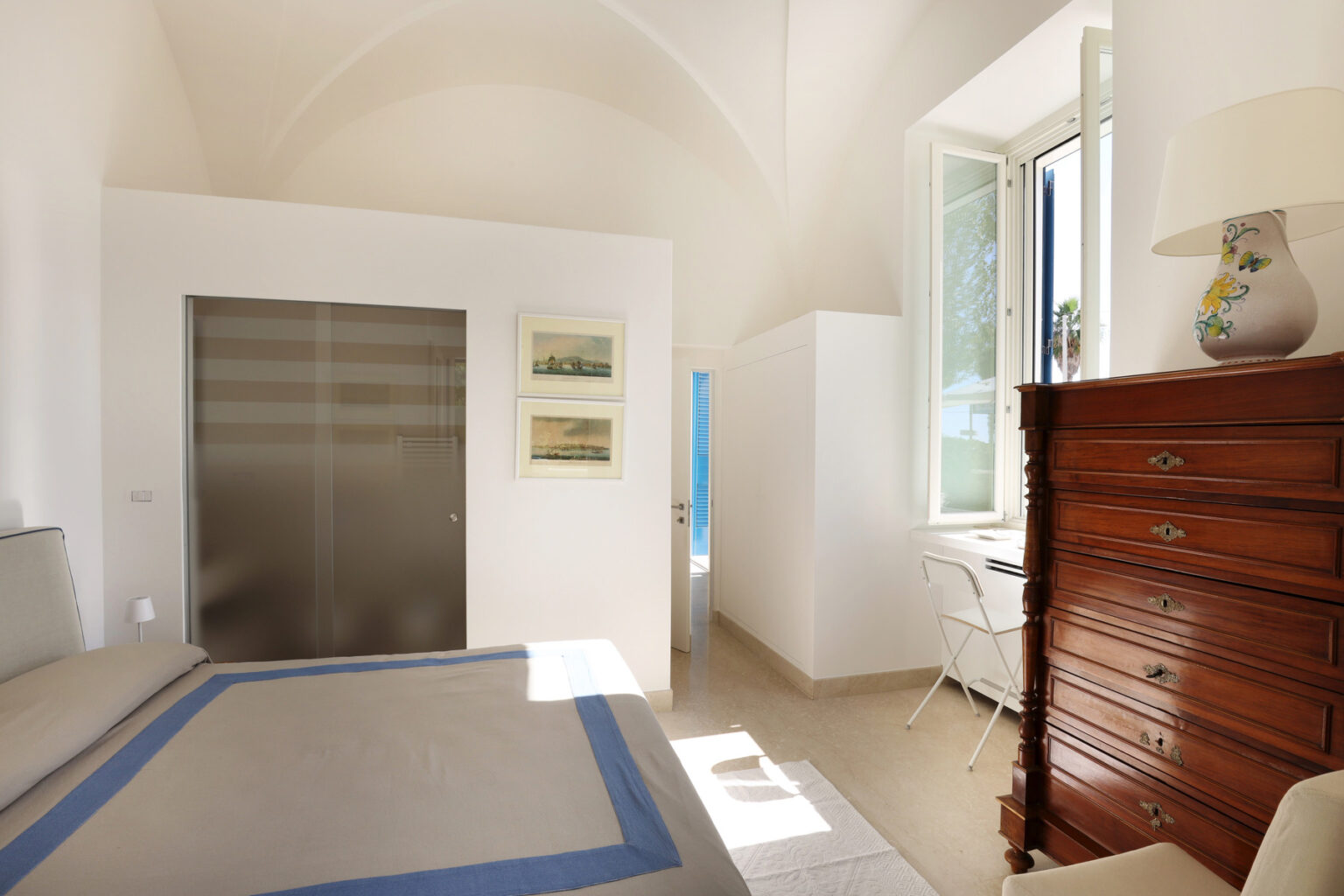 Bright and airy bedroom featuring a high vaulted ceiling, a comfortable bed with blue accents, a wooden dresser, and a large window allowing natural light. The minimalist decor includes framed artwork and a small work desk, creating a serene and inviting atmosphere. | Only on SopranoVillas
