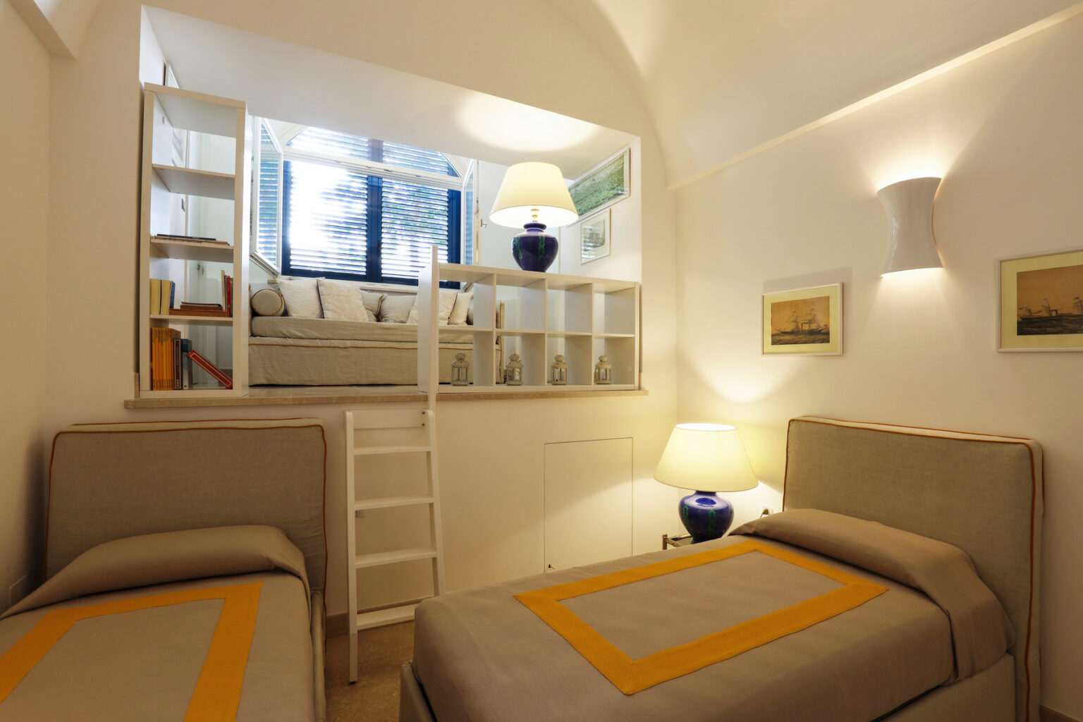 Modern bedroom interior featuring two twin beds with beige and yellow accents, a cozy reading nook with a large window, and stylish lighting. The room includes a small ladder leading to an upper level, enhancing the space's functionality and charm. | Only on SopranoVillas