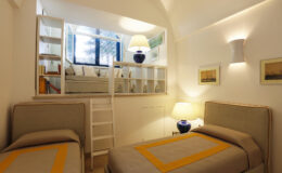Modern bedroom interior featuring two twin beds with beige and yellow accents, a cozy reading nook with a large window, and stylish lighting. The room includes a small ladder leading to an upper level, enhancing the space's functionality and charm.