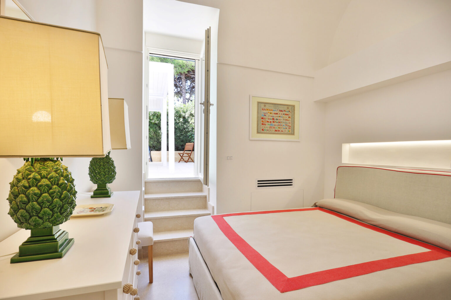 Cozy bedroom featuring a modern design with a bed adorned with a red-accented throw, a stylish lamp with a green pineapple base, and a bright, airy atmosphere created by large windows leading to an outdoor space. | Only on SopranoVillas