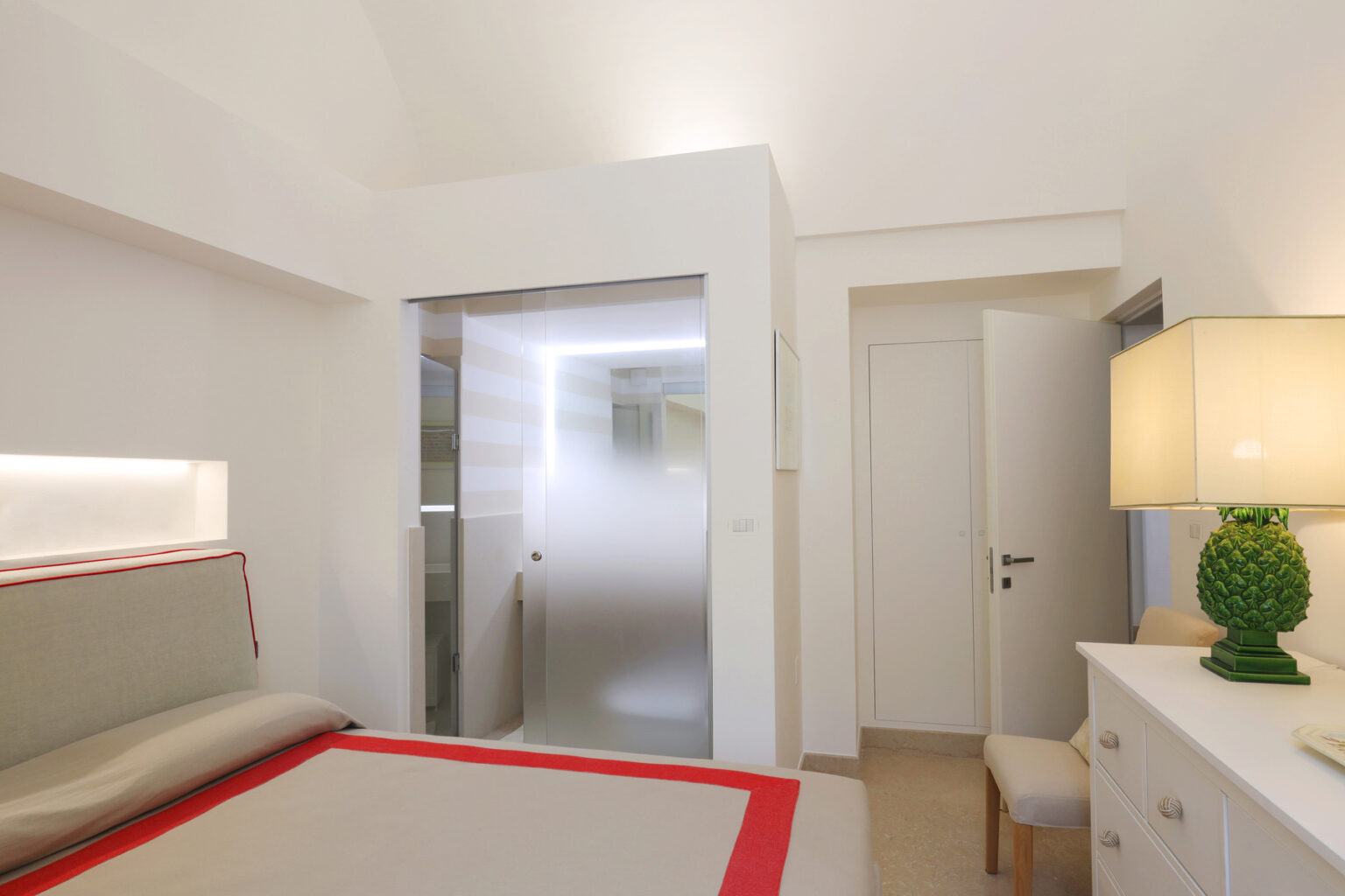 Modern bedroom interior featuring a minimalist design, a bed with a gray headboard and red accents, a frosted glass bathroom door, and a stylish lamp on a white dresser. The room is well-lit and showcases a clean, contemporary aesthetic. | Only on SopranoVillas