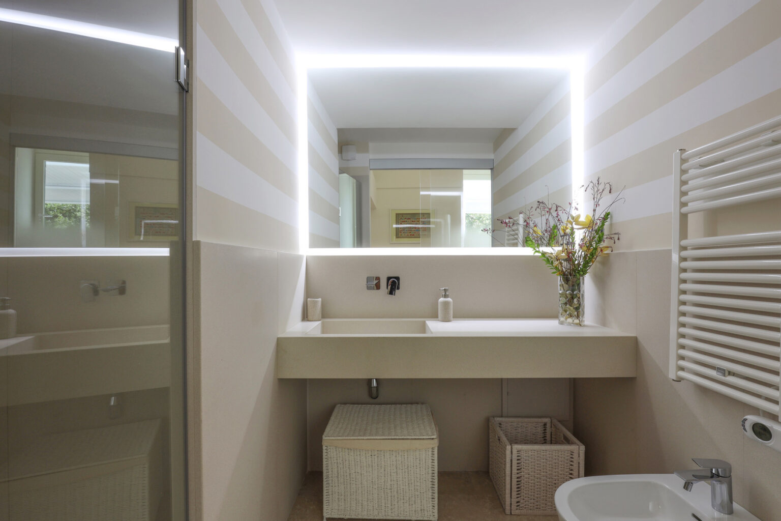 Modern bathroom interior featuring a sleek vanity with a backlit mirror, stylish storage baskets, and decorative flowers. The walls are adorned with soft striped wallpaper, enhancing the contemporary design. | Only on SopranoVillas