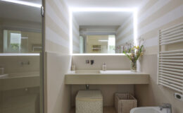 Modern bathroom interior featuring a sleek vanity with a backlit mirror, stylish storage baskets, and decorative flowers. The walls are adorned with soft striped wallpaper, enhancing the contemporary design.