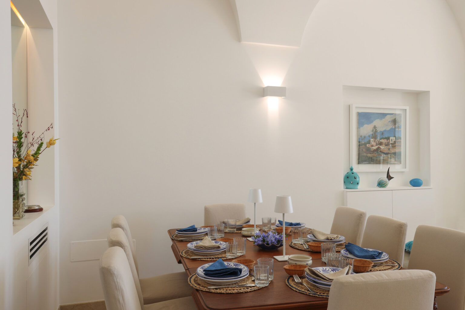 Modern dining room featuring a wooden table set for six with elegant tableware, blue napkins, and decorative accents. The bright, airy space includes white walls, soft lighting, and a framed artwork. | Only on SopranoVillas