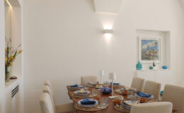 Modern dining room featuring a wooden table set for six with elegant tableware, blue napkins, and decorative accents. The bright, airy space includes white walls, soft lighting, and a framed artwork.