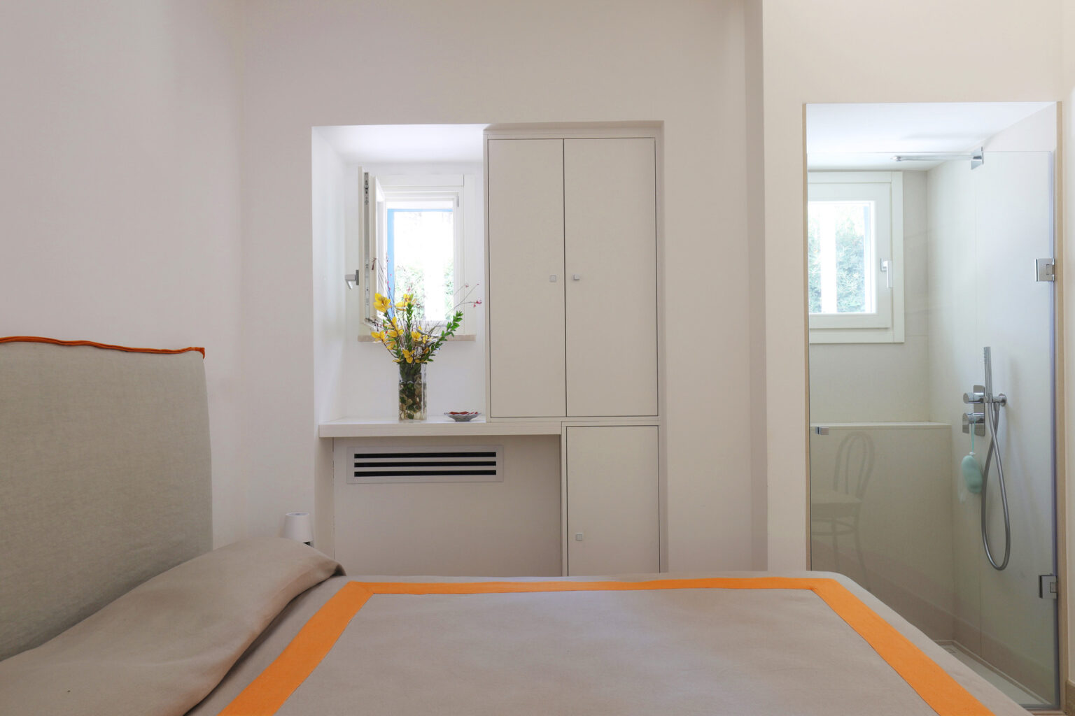 Modern bedroom featuring a light-colored bed with an orange accent, a small window with natural light, a vase of flowers on a shelf, and an adjoining bathroom with a shower. | Only on SopranoVillas