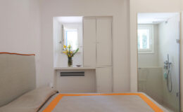 Modern bedroom featuring a light-colored bed with an orange accent, a small window with natural light, a vase of flowers on a shelf, and an adjoining bathroom with a shower.