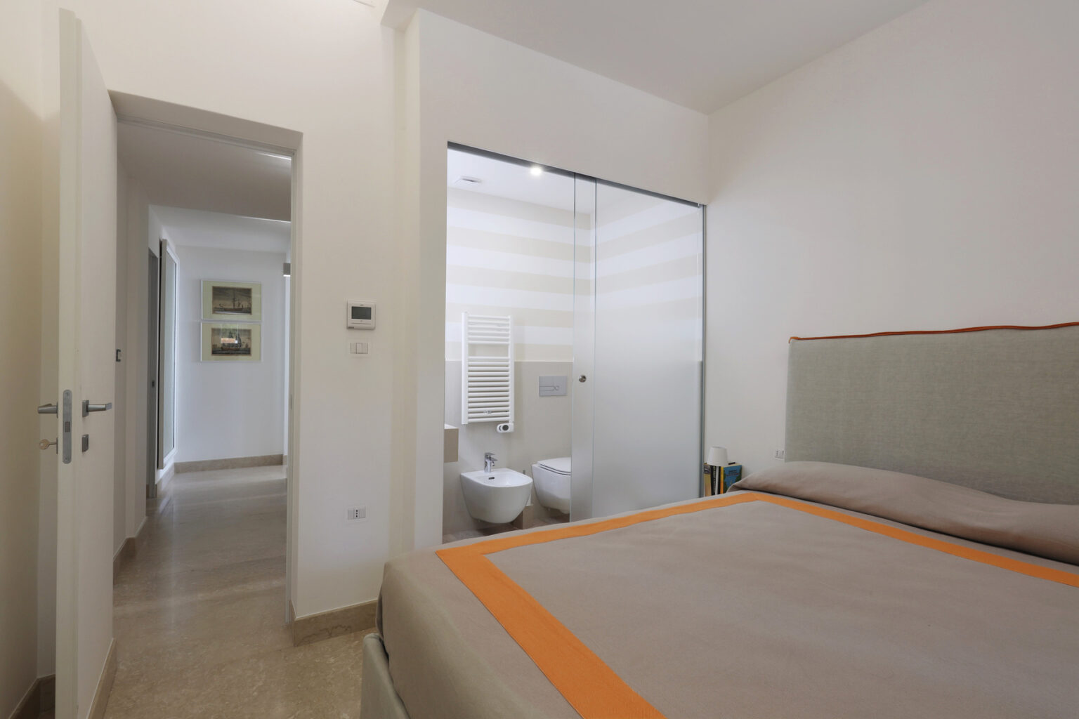 Modern bedroom with a large bed and a glass-enclosed bathroom visible in the background, featuring neutral colors and a minimalist design. The hallway leads to additional rooms, enhancing the spacious feel of the area. | Only on SopranoVillas