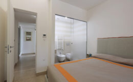 Modern bedroom with a large bed and a glass-enclosed bathroom visible in the background, featuring neutral colors and a minimalist design. The hallway leads to additional rooms, enhancing the spacious feel of the area.
