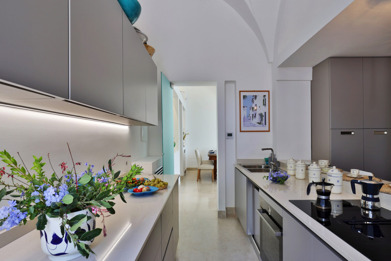 Modern kitchen interior with sleek gray cabinets, a white countertop, and a stylish coffee maker. Fresh flowers add a pop of color, while a dining area is visible in the background. Natural light enhances the inviting atmosphere. | Only on SopranoVillas