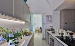 Modern kitchen interior with sleek gray cabinets, a white countertop, and a stylish coffee maker. Fresh flowers add a pop of color, while a dining area is visible in the background. Natural light enhances the inviting atmosphere.