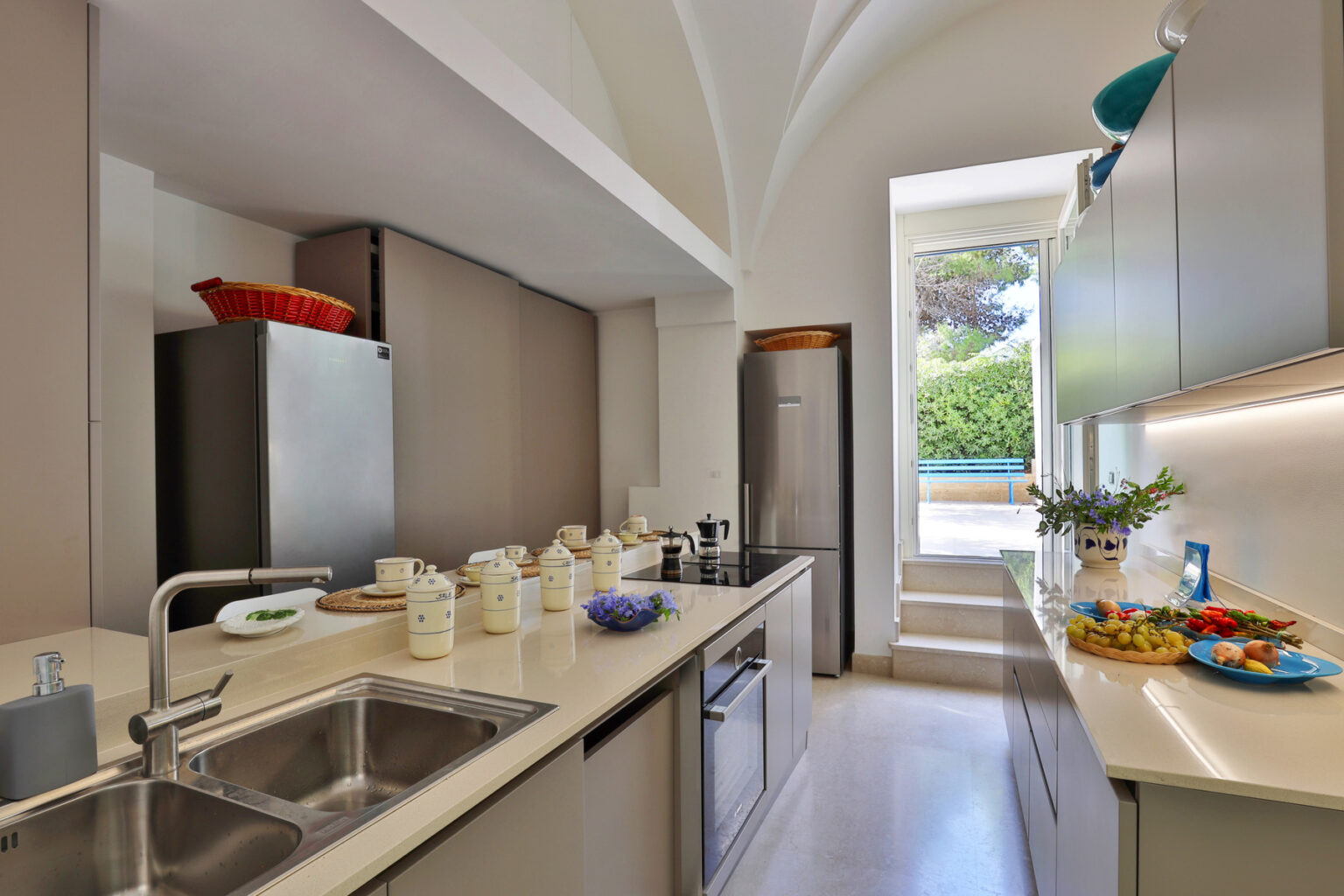 Modern kitchen interior featuring sleek cabinets, stainless steel appliances, and a bright, airy atmosphere. Natural light streams in from a large window, illuminating a countertop with decorative bowls and jars. | Only on SopranoVillas