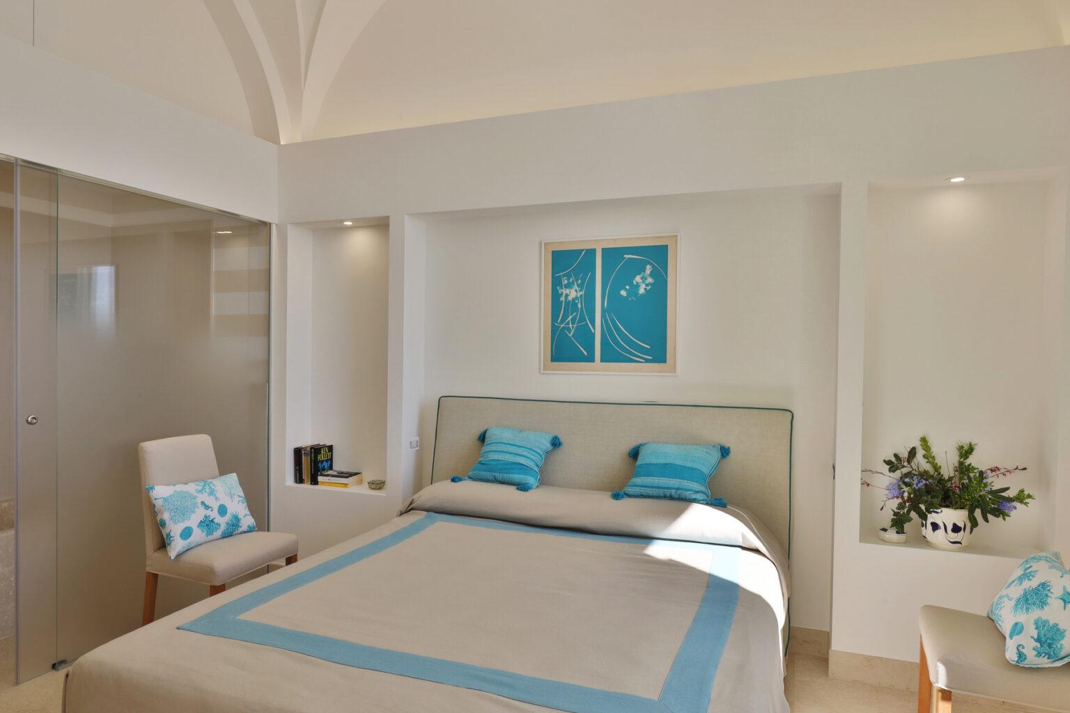 Modern bedroom interior featuring a light color palette with blue accents, a comfortable bed with decorative pillows, and stylish decor elements. The room includes a cozy chair and a small plant, enhancing the inviting ambiance. | Only on SopranoVillas