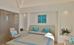 Modern bedroom interior featuring a light color palette with blue accents, a comfortable bed with decorative pillows, and stylish decor elements. The room includes a cozy chair and a small plant, enhancing the inviting ambiance.