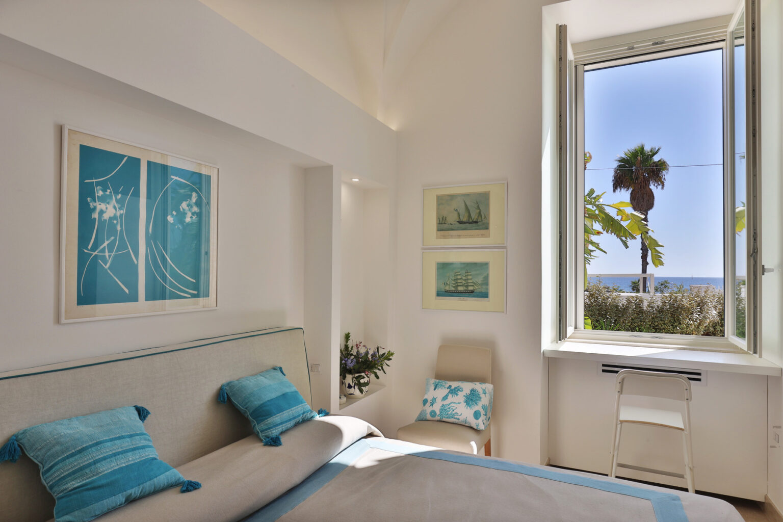 Bright and airy bedroom with a coastal theme, featuring blue accent pillows, artwork on the walls, and a large window offering a view of palm trees and the ocean. | Only on SopranoVillas