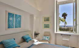 Bright and airy bedroom with a coastal theme, featuring blue accent pillows, artwork on the walls, and a large window offering a view of palm trees and the ocean.