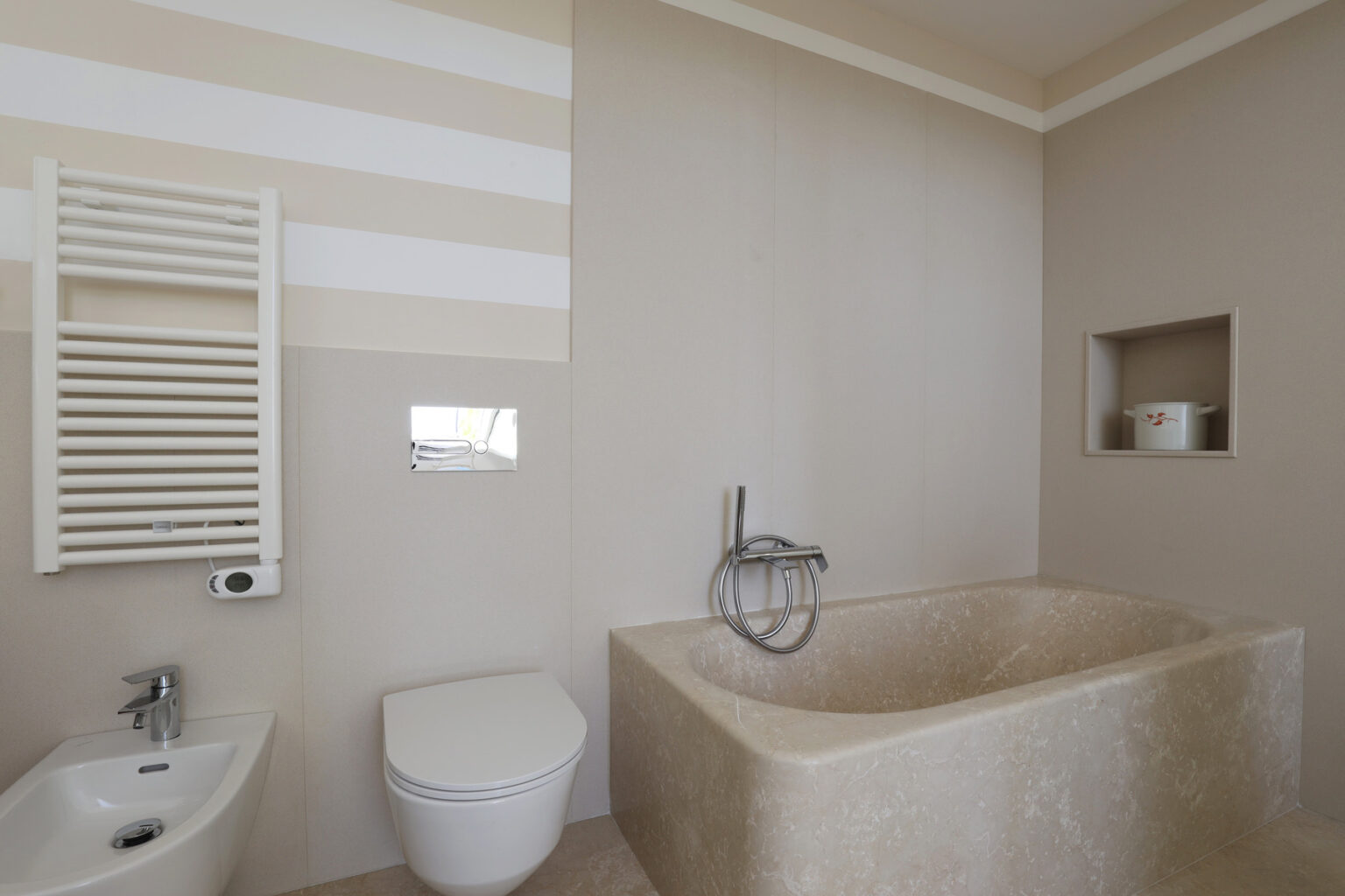 Modern bathroom featuring a minimalist design with a stone bathtub, wall-mounted faucet, and a toilet. The walls are painted in soft colors with horizontal stripes, complemented by a towel radiator and a recessed shelf for storage. | Only on SopranoVillas