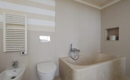 Modern bathroom featuring a minimalist design with a stone bathtub, wall-mounted faucet, and a toilet. The walls are painted in soft colors with horizontal stripes, complemented by a towel radiator and a recessed shelf for storage.