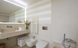 Modern bathroom interior featuring a stylish sink, toilet, bidet, and a mirror with ambient lighting. The walls are adorned with soft striped patterns, and a decorative plant adds a touch of elegance to the space.