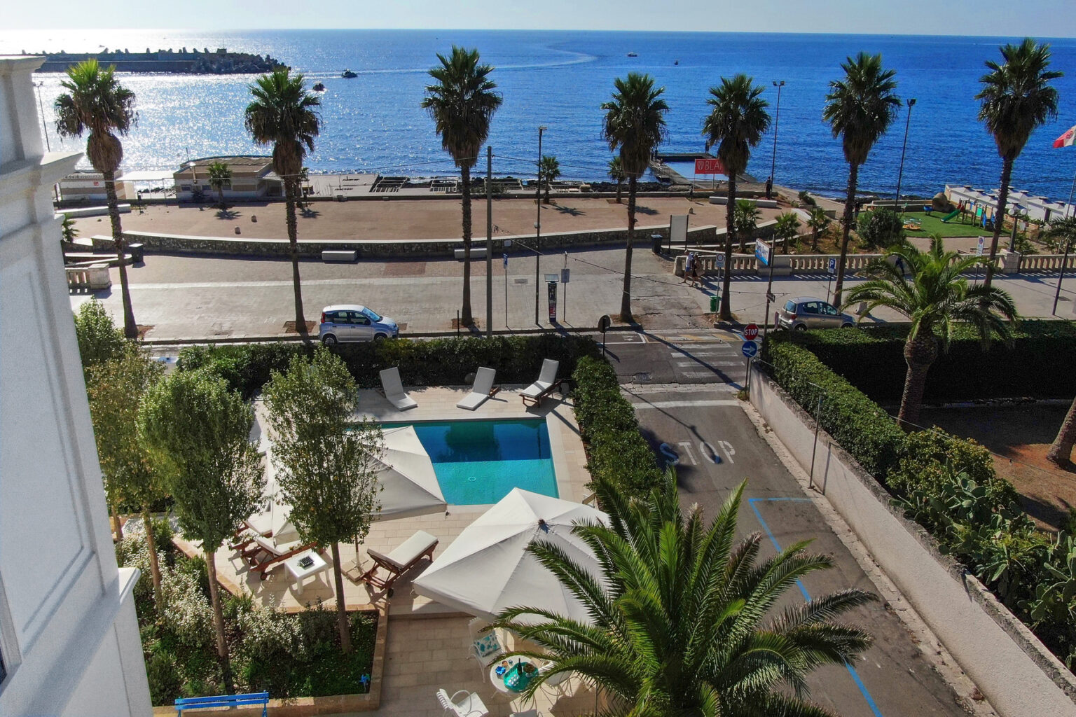View of a seaside hotel pool surrounded by palm trees, with a beach and ocean in the background, showcasing a sunny day and inviting atmosphere. | Only on SopranoVillas