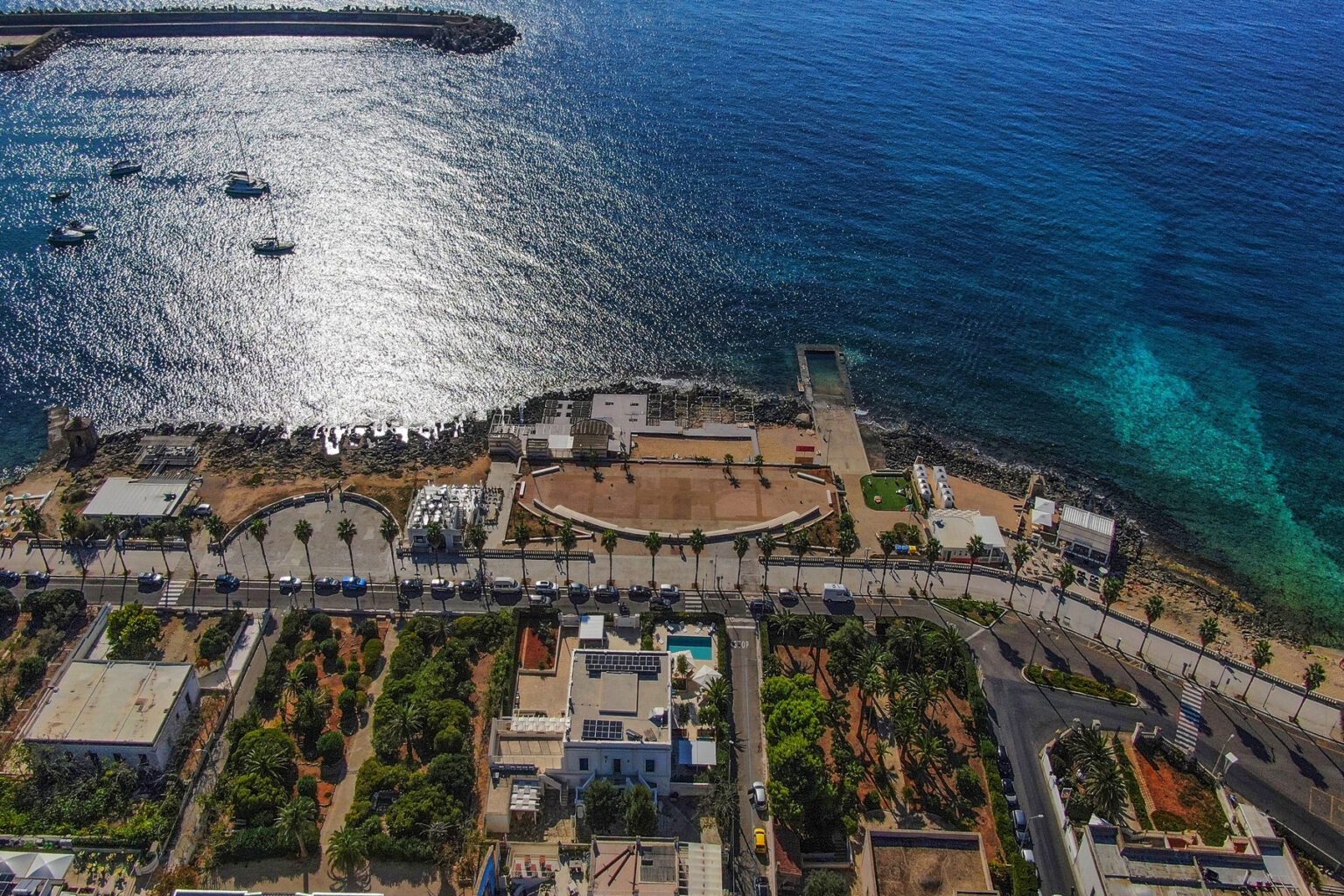 Aerial view of a coastal promenade with lush gardens and a clear blue sea, featuring boats in the water and a scenic shoreline. Ideal for showcasing waterfront living and tourism in a vibrant seaside location. | Only on SopranoVillas