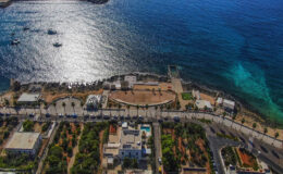 Aerial view of a coastal promenade with lush gardens and a clear blue sea, featuring boats in the water and a scenic shoreline. Ideal for showcasing waterfront living and tourism in a vibrant seaside location.