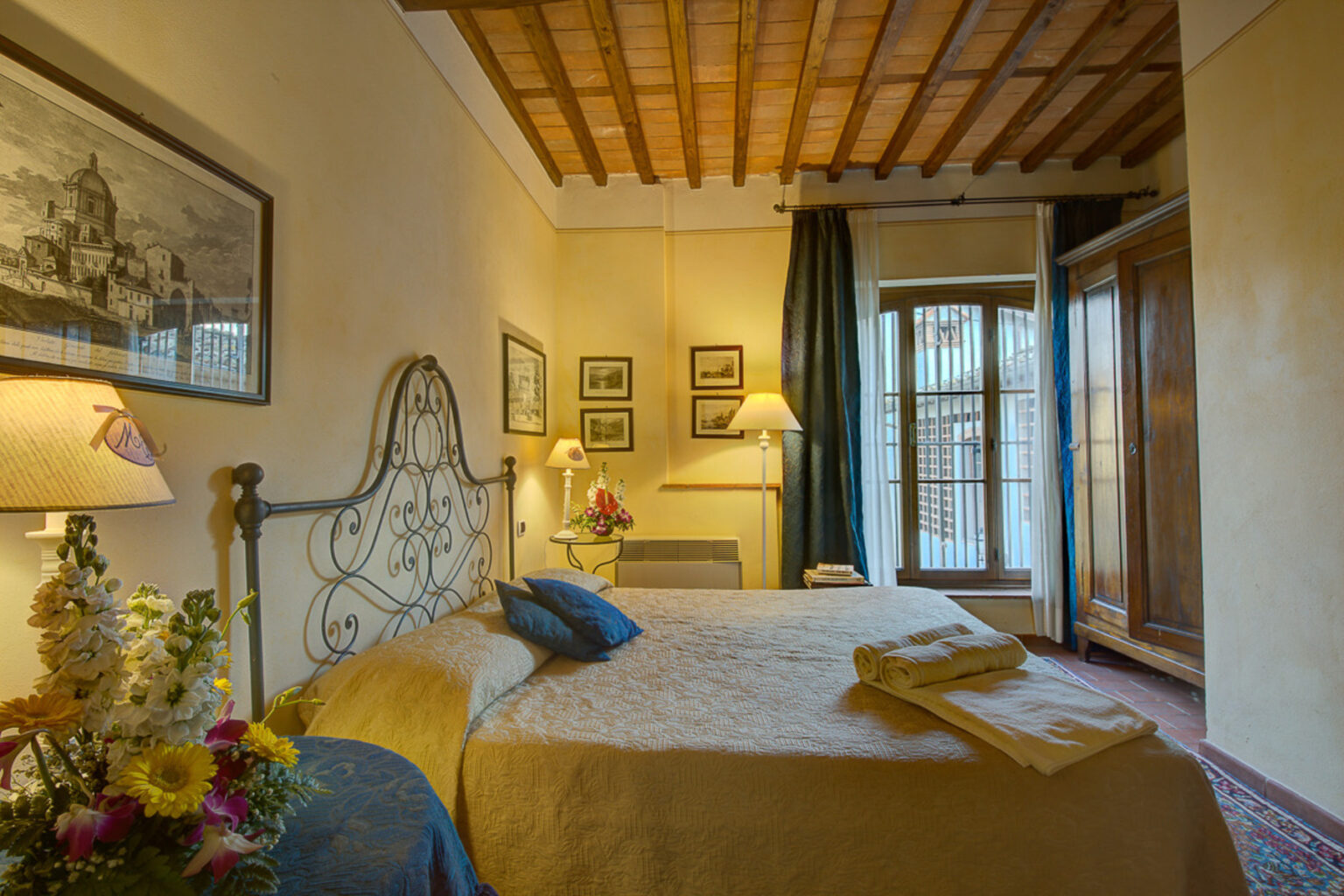 Cozy bedroom featuring a wrought iron bed, warm yellow walls, and wooden ceiling beams. The room includes decorative artwork, bedside lamps, and a view of a balcony with curtains. Fresh flowers add a touch of elegance, creating a welcoming atmosphere for guests. | Only on SopranoVillas