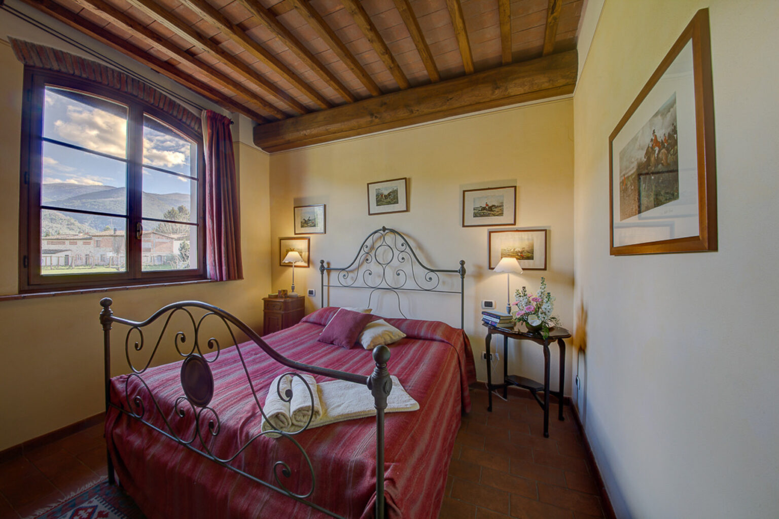 Cozy bedroom featuring a wrought iron bed with red bedding, wooden ceiling beams, and a large window allowing natural light. The room includes bedside lamps, decorative artwork on the walls, and a small table with a floral arrangement, creating an inviting atmosphere. | Only on SopranoVillas