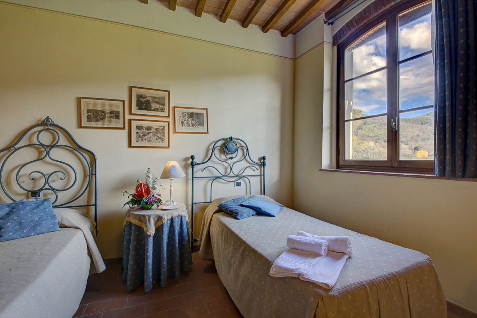 Cozy guest room featuring two twin beds with decorative pillows, a bedside table with a lamp, and framed artwork on the walls, illuminated by natural light from a large window with scenic views. | Only on SopranoVillas