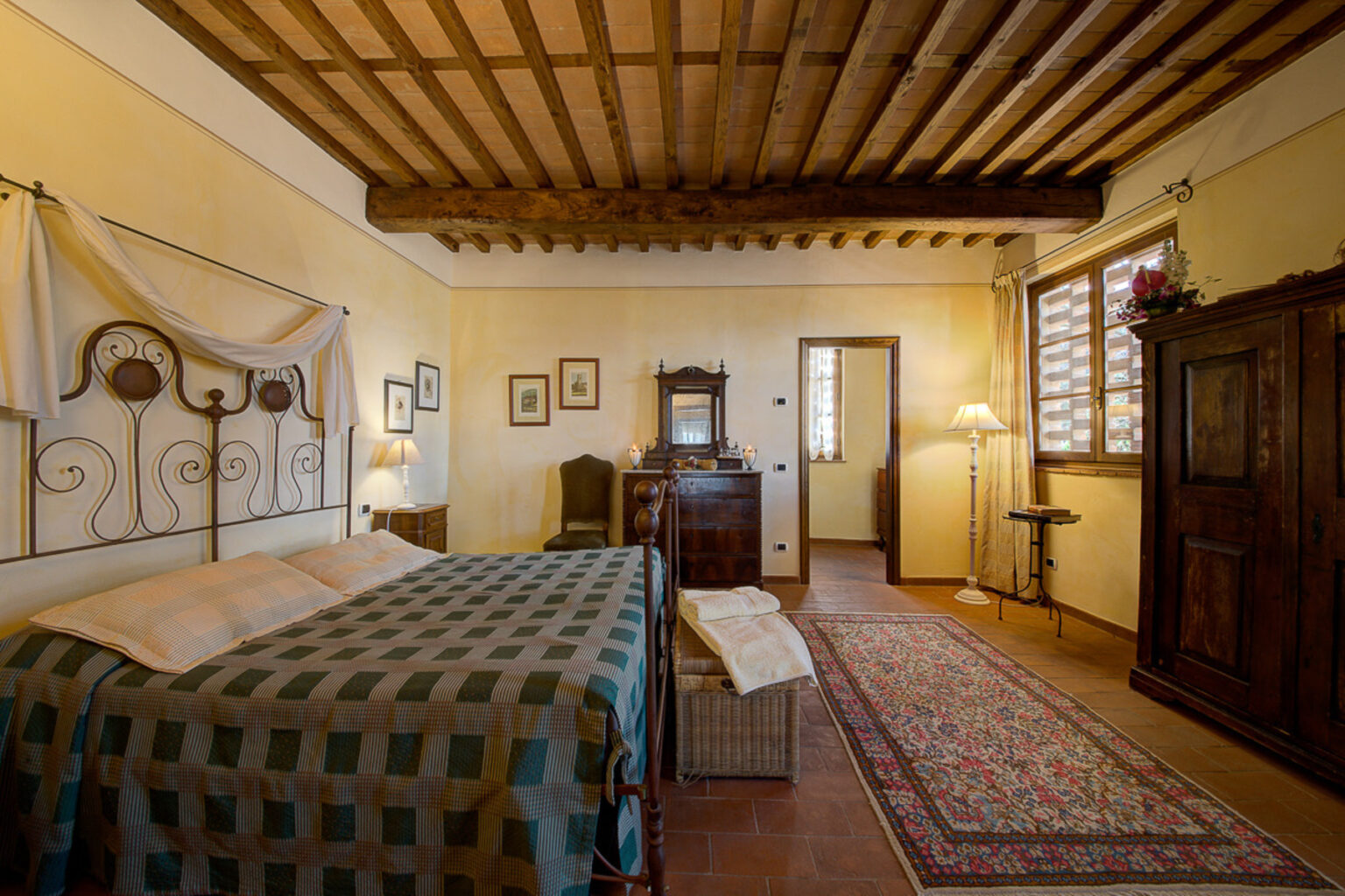 Cozy bedroom featuring a wrought iron bed with a patterned quilt, wooden ceiling beams, and warm yellow walls. The room includes a vintage dresser, bedside lamp, and decorative art on the walls, creating a charming and inviting atmosphere. | Only on SopranoVillas