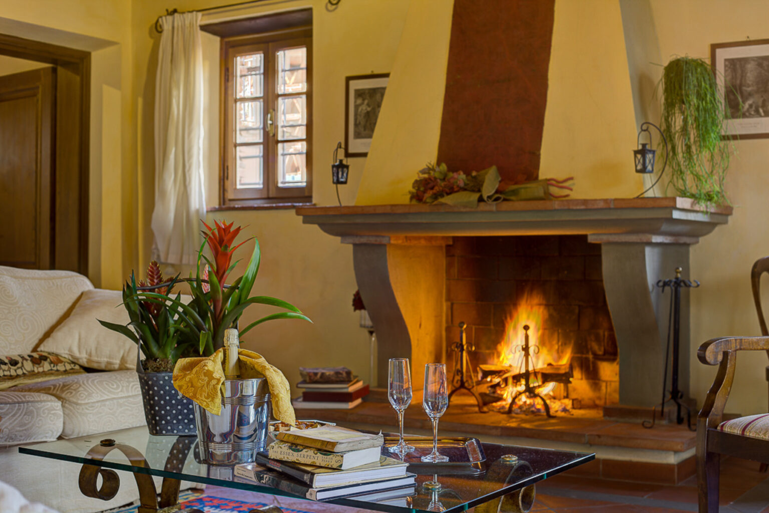 Cozy living room featuring a stylish glass coffee table with books and champagne flutes, a vibrant potted plant, and a warm fireplace with a rustic mantel, surrounded by natural light from windows adorned with white curtains. | Only on SopranoVillas