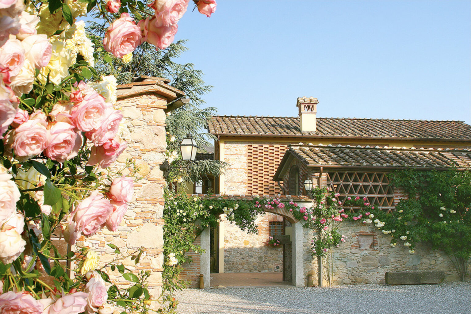 Beautiful rustic villa surrounded by blooming pink and white roses, featuring stone walls and a tiled roof under a clear blue sky. Ideal for showcasing charming countryside architecture and vibrant gardens. | Only on SopranoVillas