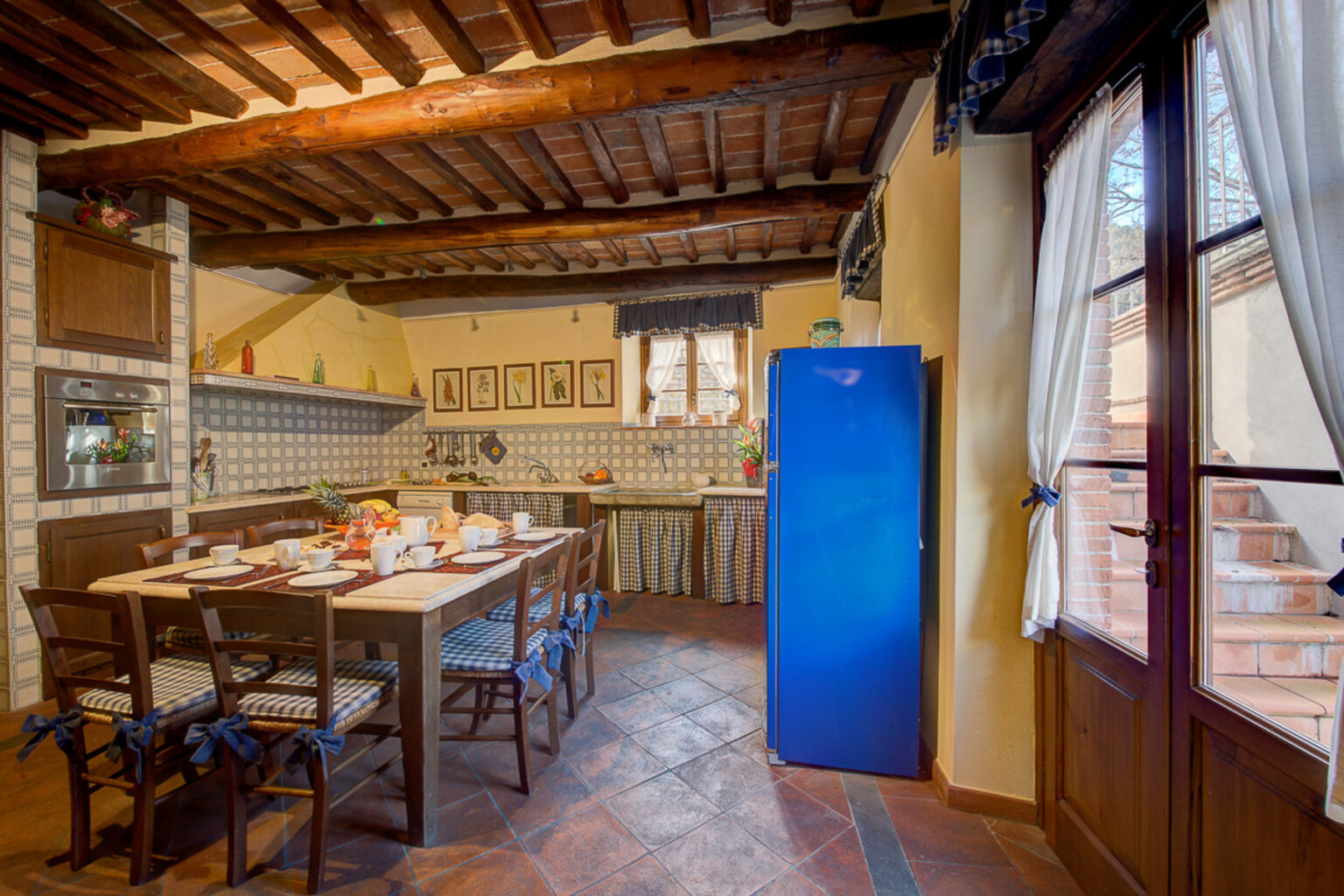 Cozy rustic kitchen featuring wooden beams, tiled backsplash, and a blue refrigerator. A wooden dining table is set for eight, surrounded by chairs with blue ribbon accents. Sunlight streams through the window, highlighting the charming decor and inviting atmosphere. | Only on SopranoVillas
